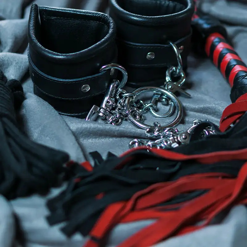 Black leather bondage gear with cuffs, chain links, and a red and black flogger