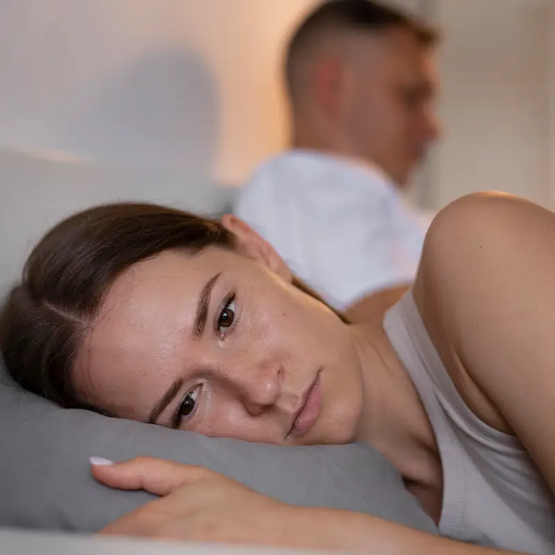 Woman lying awake while partner faces away, reflecting emotional distance and intimacy struggles
